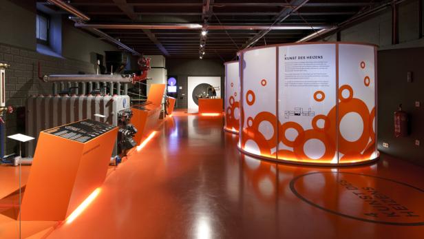 Innenansicht eines Museumsraums mit orangefarbenem Boden und Ausstellungsstücken zum Thema Heizen.
