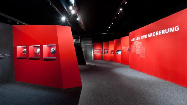 Innenansicht einer Ausstellung mit dem Titel „Wellen der Eroberung“ und antiken Masken in Vitrinen.