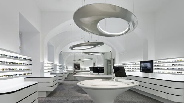 Innenansicht eines Optikergeschäfts mit Brillen in Regalen und runden Tischen.