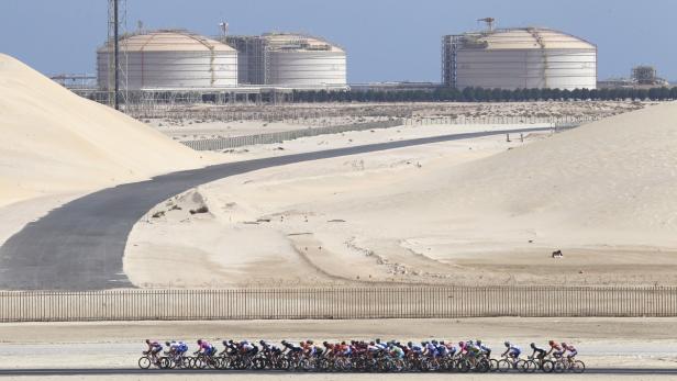 Eine Gruppe Radfahrer fährt auf einer Straße in der Wüste, im Hintergrund große Tanks.