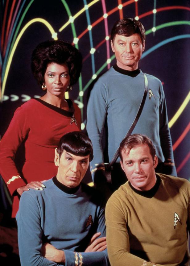 Ein Gruppenbild der Besetzung von „Star Trek“ in ihren Uniformen vor einem bunten Hintergrund.