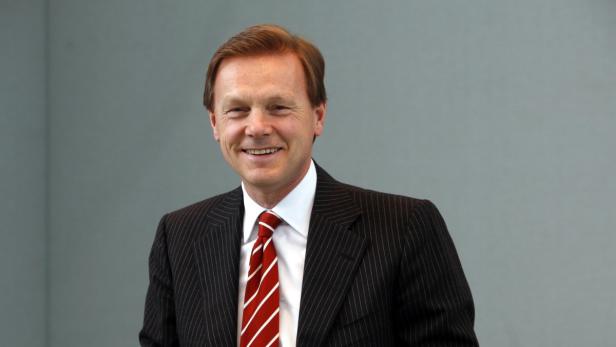 Ein lächelnder Mann im Anzug mit rot-gestreifter Krawatte vor einem grauen Hintergrund.