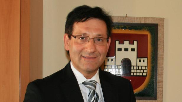 Ein Mann mit Brille und Anzug vor einem Wappenbild.