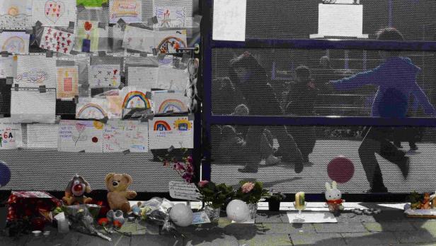 Vor einem Gitter hängen Kinderzeichnungen und liegen Blumen, Stofftiere und Kerzen.