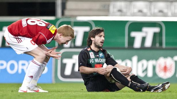 Ein Fußballspieler sitzt verletzt auf dem Rasen, während ein anderer sich aufregt.