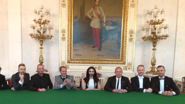 Conchita Wurst sitzt mit anderen Personen an einem Tisch vor einem Gemälde.