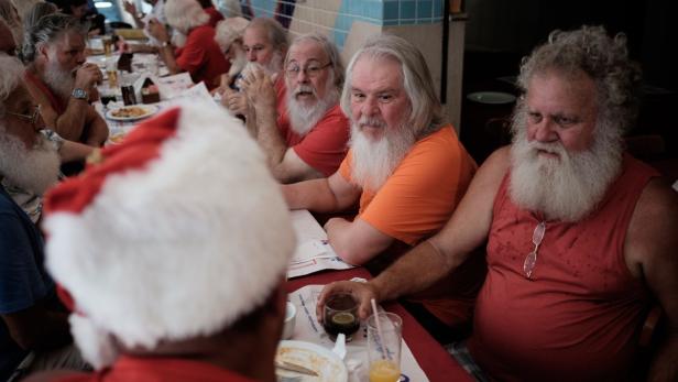 Mehrere Männer mit weißen Bärten sitzen an einem Tisch, einige tragen Weihnachtsmannmützen.