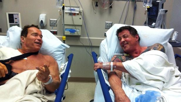 Arnold Schwarzenegger und Sylvester Stallone liegen nebeneinander in Krankenhausbetten und unterhalten sich.