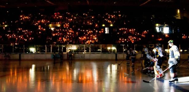 Eishockeyspieler auf dem Eis, während die Zuschauer im Hintergrund Kerzen halten.