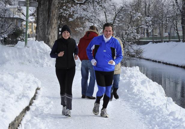 Eine Frau und ein Mann joggen im Winter an einem Fluss entlang.
