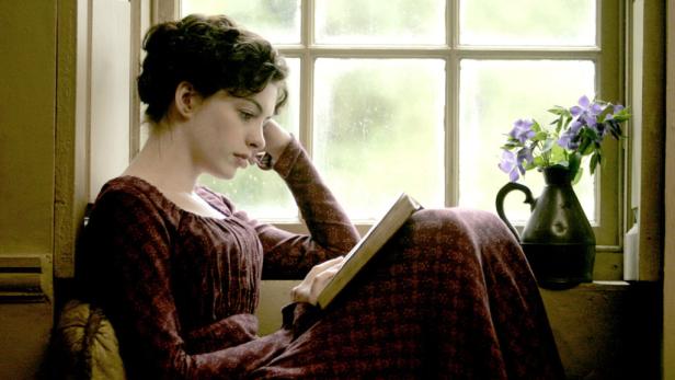 Eine junge Frau im Regency-Kleid liest am Fenster ein Buch.