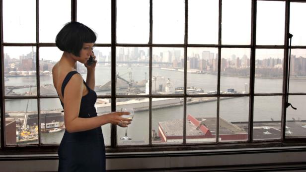 Eine Frau telefoniert mit einem Glas Wein in der Hand vor einem Fenster mit Blick auf die Skyline.