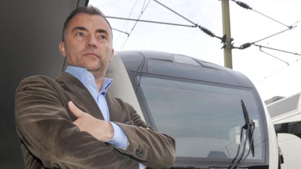 Ein Mann in einem braunen Sakko steht vor einem modernen Zug.