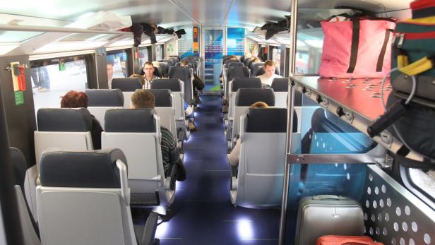 Blick in einen modernen Zugwagen mit Fahrgästen und Gepäckablagen.