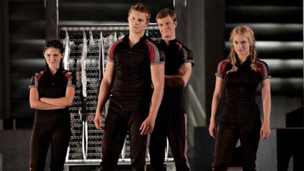 Vier Schauspieler in Kostümen von „Die Tribute von Panem“ posieren vor einem Gitter.