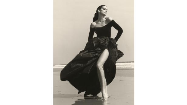 Eine Frau in einem schwarzen Kleid mit Beinschlitz steht am Strand.
