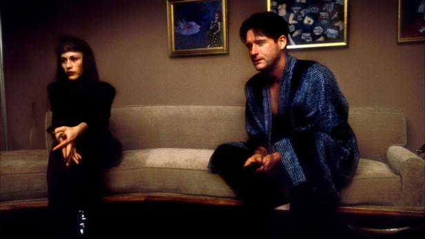 Ein Mann im Morgenmantel und eine Frau in Schwarz sitzen auf einem Sofa.