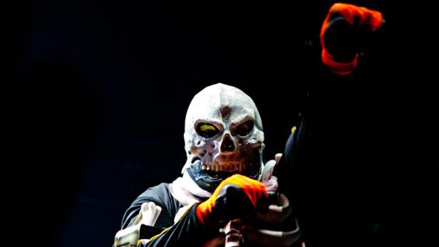 Eine Person mit einer Totenkopfmaske und orangefarbenen Handschuhen vor dunklem Hintergrund.