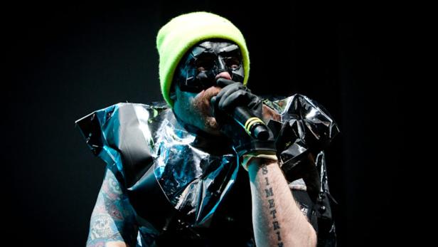 Ein maskierter Sänger mit neonfarbener Mütze tritt auf einer Bühne auf.