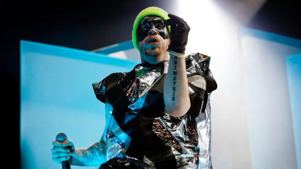 Ein Mann mit neonfarbener Mütze und Maske hält ein Mikrofon und trägt ein ungewöhnliches Kostüm.