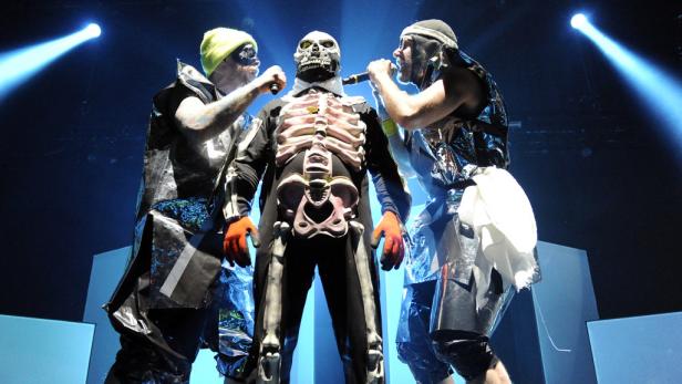 Drei Performer in ausgefallenen Kostümen stehen auf einer Bühne und singen.