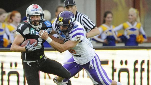 Zwei American-Football-Spieler kämpfen um den Ball, während Cheerleader im Hintergrund zu sehen sind.