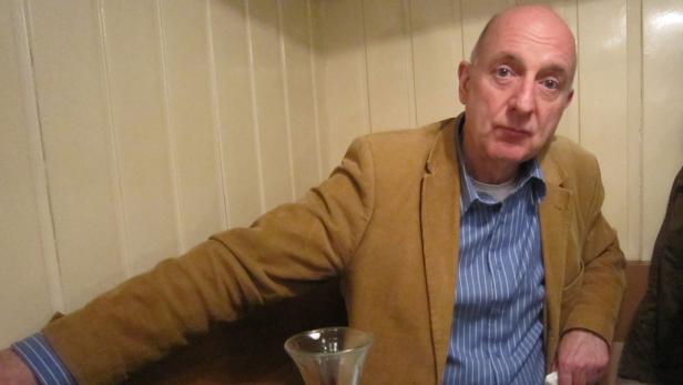 Ein Mann mit hellbraunem Sakko und blau gestreiftem Hemd sitzt an einem Tisch.