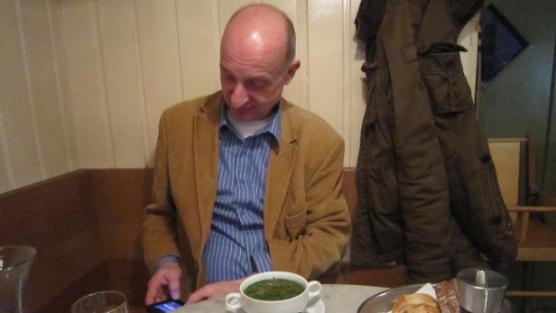 Ein Mann sitzt an einem Tisch, blickt auf sein Smartphone und isst eine Suppe.