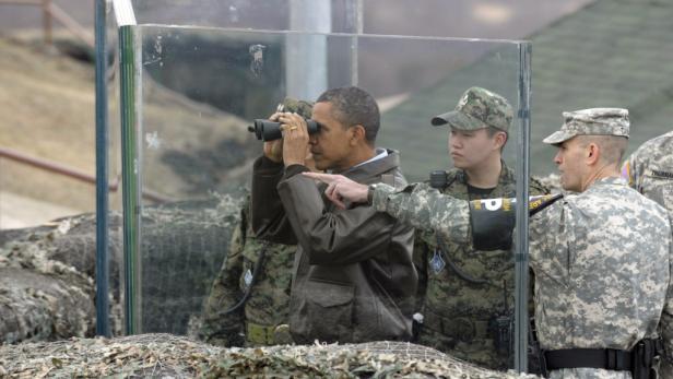 Barack Obama blickt durch ein Fernglas hinter einer Glasscheibe, umgeben von Soldaten.