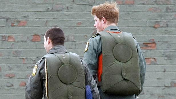 Prinz Harry und ein weiterer Mann in Uniform und mit Schwimmwesten vor einer Ziegelwand.