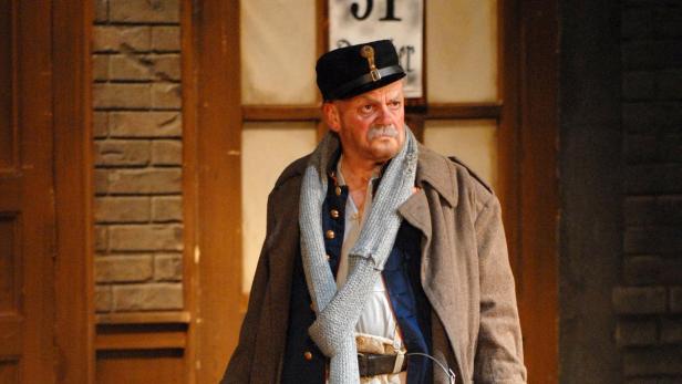 Ein Mann mit Schnurrbart, Uniform und Mantel steht vor einer Backsteinwand.