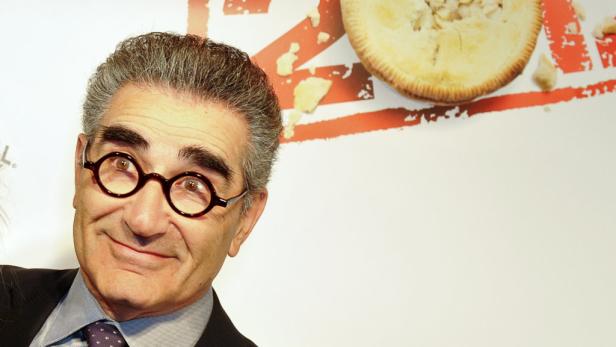Eugene Levy mit runder Brille vor einem Hintergrund mit einem Kuchen.