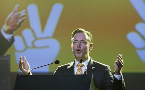 Ein Mann in Anzug gestikuliert vor einem Hintergrund mit einem Friedenszeichen.