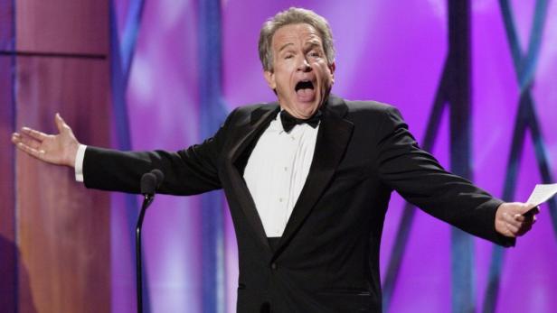 Ein Mann im Smoking gestikuliert auf einer Bühne mit offenem Mund.