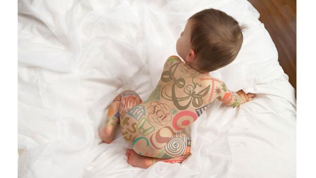 Ein Baby liegt auf dem Bauch auf einem weißen Laken und trägt einen Ganzkörperanzug mit bunten Mustern.