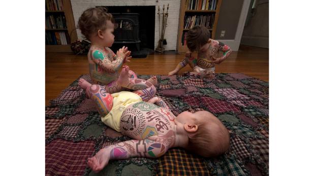Drei Babys mit aufgemalten Tattoos liegen auf einer bunten Patchworkdecke.