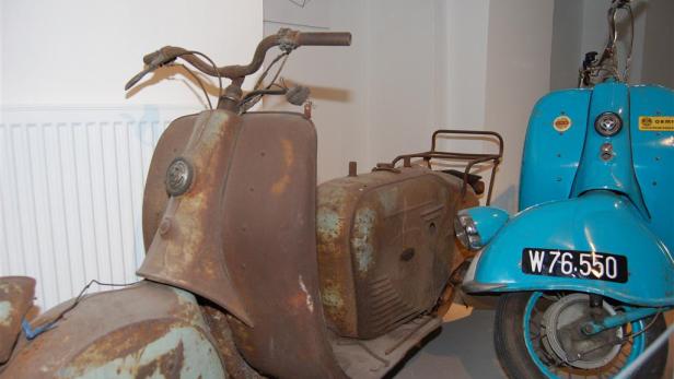 Ein verrosteter Motorroller steht neben einem türkisfarbenen Roller mit dem Kennzeichen W 76.550.