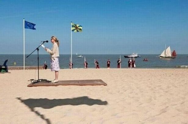 Eine Frau hält eine Rede am Strand, im Hintergrund Flaggen und eine Ehrenformation.