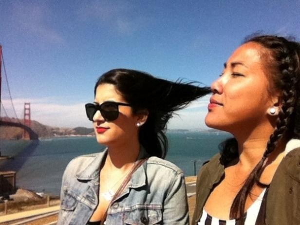 Zwei Frauen posieren vor der Golden Gate Bridge, wobei der Wind die Haare der einen ins Gesicht der anderen weht.