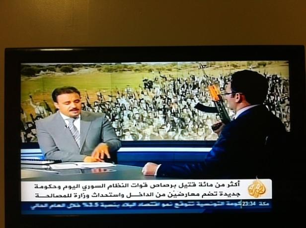 Zwei Männer sitzen in einem Fernsehstudio vor einem Bildschirm mit einer Menschenmenge und Gewehren.