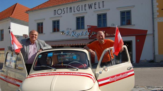 Zwei Männer posieren vor einem Oldtimer-Fiat mit österreichischen Flaggen vor dem Geschäft „Nostalgiewelt“.