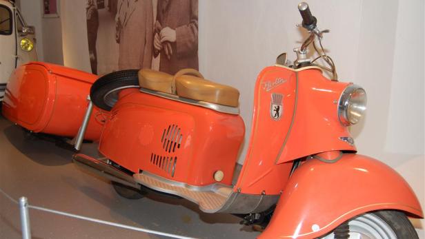 Ein orangefarbener Roller „Berlin“ mit Anhänger steht in einem Museum.