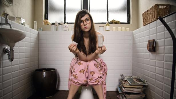 Eine Frau mit Brille sitzt auf der Toilette und hält eine leere Toilettenpapierrolle und einen Spender in den Händen.