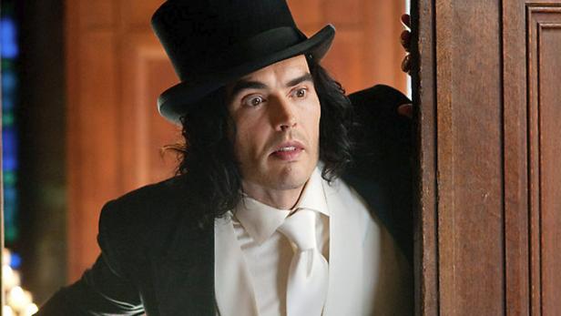 Russell Brand mit Zylinder schaut hinter einer Holztür hervor.