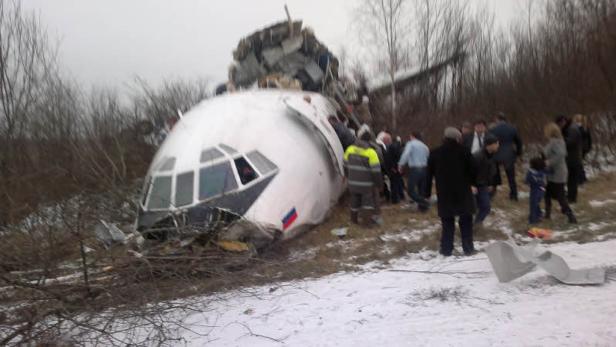 Ein Flugzeugwrack liegt in einem verschneiten Feld, umgeben von Menschen.