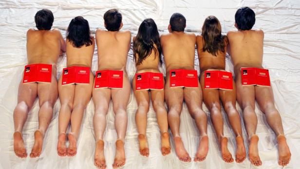 Sieben nackte Personen liegen auf dem Bauch, jeweils mit einem roten Buch auf dem unteren Rücken.