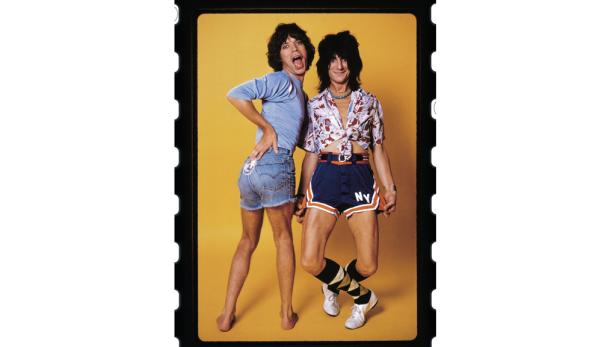 Mick Jagger und Ron Wood posieren vor einem gelben Hintergrund.