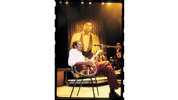 Chuck Berry sitzt auf einem Stuhl und spielt Gitarre vor einer großen Leinwand.