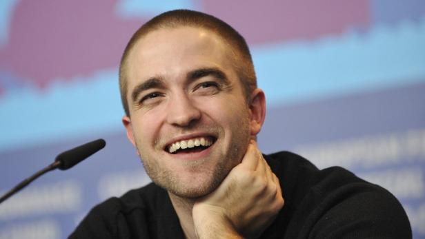 Robert Pattinson lächelt mit der Hand am Kinn in die Kamera.