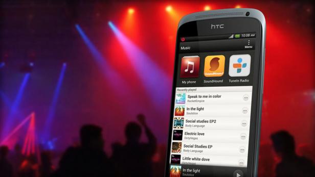 Ein HTC-Smartphone zeigt eine Musik-App vor dem Hintergrund einer Menschenmenge mit Bühnenbeleuchtung.